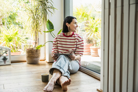 Smiling woman holding tablet PC and looking out of window at home