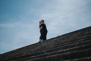A scene capturing two individuals hugging on cement steps with a serene sky above. The minimalistic composition emphasizes connection, comfort, and an urban environment blending with nature.