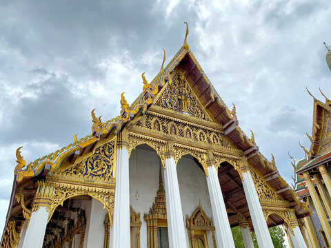 Magnificent golden temple architecture at the Grand Palace, Bangkok. Intricate Thai traditional carvings and gables under a dramatic cloudy sky. Worshipper area view.