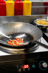 Stir-frying sliced bell peppers and onions in a wok on an induction cooktop. Noodles in a bowl nearby. Professional kitchen scene with vibrant ingredients and cooking tools