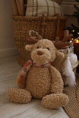 A soft, fluffy stuffed reindeer sits on a warm floor. It has a scarf and antlers, adding a festive touch to the cozy setting filled with holiday decor.