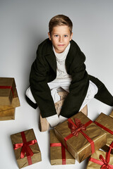 Cute preteen boy in stylish winter outfit surrounded by wrapped gifts