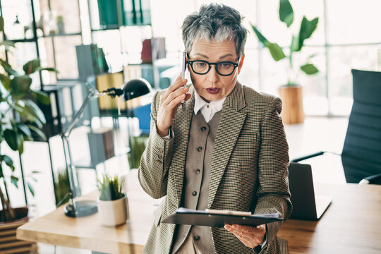 Professional businesswoman in elegant formalwear talking on phone in modern office environment with digital tablet - Powered by Adobe