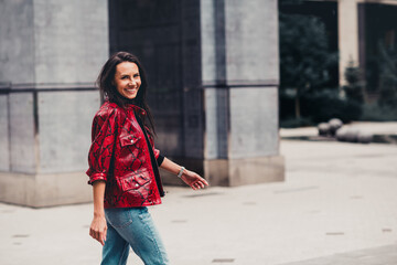 Fototapeta premium Beautiful young woman in a red jacket walking outdoors in a city during daylight, showcasing casual urban fashion
