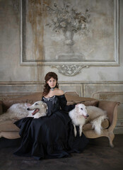 An elegant dark-haired girl with two greyhounds is sitting on a sofa in a vintage interior