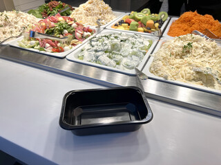 A self-service buffet counter with fresh salads, fruit, and vegetables displayed in trays with an empty black container in the foreground