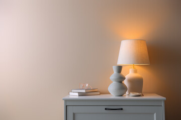 Stylish lamp, vase, pearl bracelet, books and glasses on white dresser near beige wall indoors, space for text