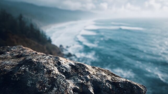 A rugged rocky cliff edge overlooks a vast misty ocean with rolling waves towards a distant shoreline and forested hills
