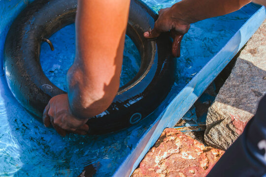 A person's hands meticulously inspect a bicycle tire inner tube in a water basin to locate a puncture at a tire repair shop in India, captured from an overhead perspective.