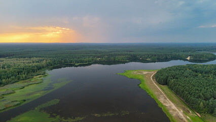 Obraz premium Aerial view of a serene lake surrounded by lush green forests at sunset, highlighting the tranquil beauty of nature. Ideal for nature enthusiasts and outdoor activities.