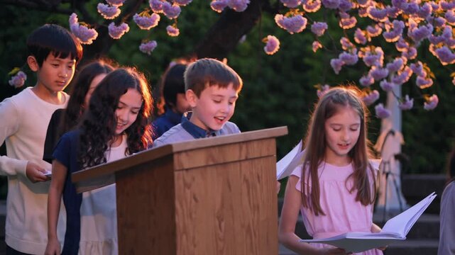 Group of diverse elementary school children performing a song during a school event, singing happily in a chorus from a wooden lectern with cherry blossoms in the background