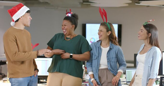 Diverse coworkers laughing, adjusting headbands in office after colleague in Santa hat speaking