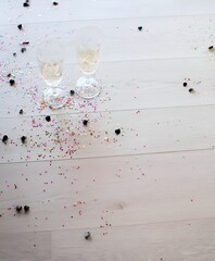 Two elegant champagne glasses standing on a light wooden floor surrounded by colorful confetti and black decorative pieces after a celebration. Bright soft daylight and festive mood.