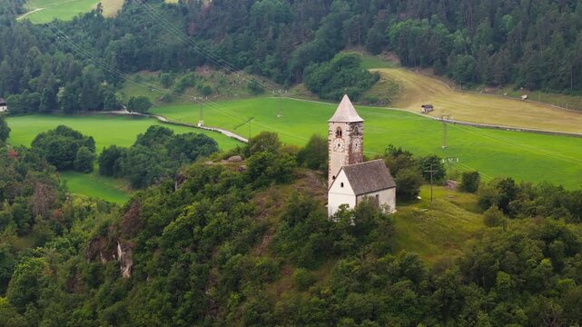 Chiesa di Santa Verena crowning a forested hill near a valley edge, aerial view. Northern Italy mountain landscape