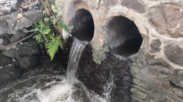 Water flows from two concrete pipes embedded in the stone wall. The water flows into a small stream below.