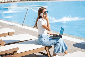 a businesswoman is working on a laptop and talking on the phone at a hotel poolside on a sunbed in the summer