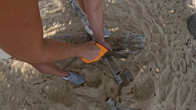 Hands Using Handsaw to Cut Wooden Log on Sandy Beach, Close Up from Overhead Perspective, Outdoor Crafting Concept
