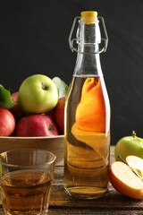 Delicious cider in bottle, glass and apples on wooden table against dark gray background, closeup