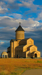 Golden-hour view of the Church of the Iveron Icon of the Mother of God in Tbilisi, Georgia, with dramatic clouds and vivid golden grass in the foreground.