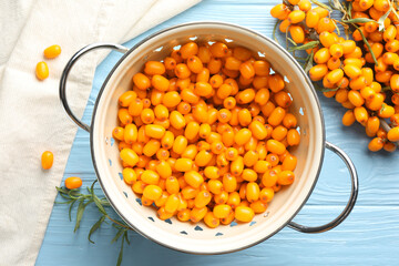 Fresh sea buckthorn berries in metal colander and leaves on light blue wooden table, flat lay