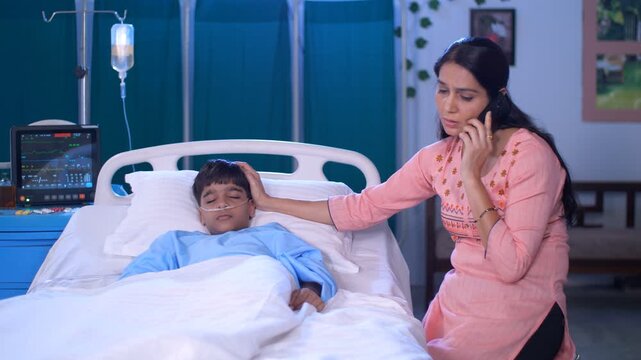 A stressed Indian woman in her late forties is talking on mobile in the hospital - caring mother  single mother  hospital care. A responsible Indian mother is by the bedside of her sick son - singl...