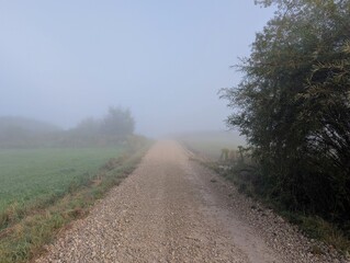 A horizontal image of a rural gravel road vanishing into dense morning fog, surrounded by green fields and vegetation. A vertical version of this photo is also available.
