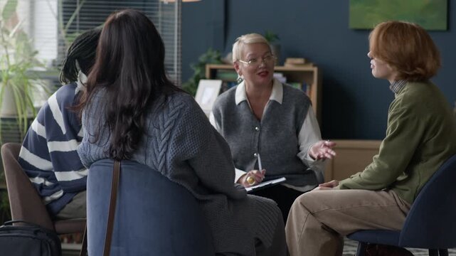 Medium long shot of middle-aged Caucasian female counselor having group therapy session with young diverse students sitting in circle in modern office and engaged in conversation