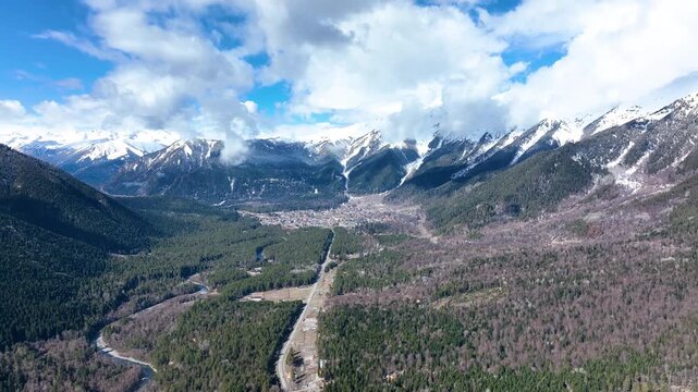 A majestic aerial panorama of a deep, wide mountain valley with a resort town nestled in the center, surrounded by dense pine forests and a backdrop of towering, snow-capped peaks under a bright, clou