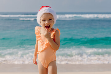 Child girl in Christmas hat on beach with tropical sea. Happy smiling girl