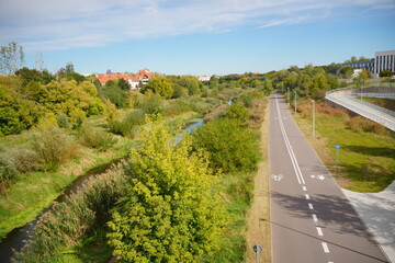 Scenic winding bicycle and walking path alongside a tranquil river, surrounded by lush green trees and vibrant flora under a clear blue sky