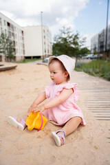 young child wearing a white sun hat and pink dress sits on sand in a residential playground, engaging with a bright yellow bucket under a clear blue sky, outdoor, sunlight, carefree, cheerful