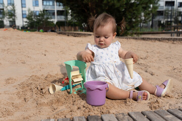 adorable child playing in urban sandbox surrounded by toy buckets and sunny weather in residential area with trees and buildings in the background, patio, sandcastles, interaction, joy, curiosity
