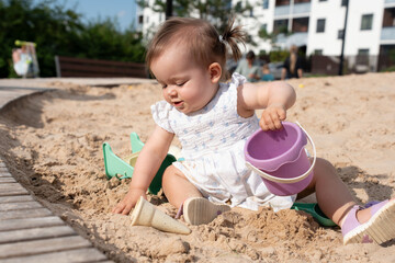 adorable toddler joyfully playing with a purple bucket in a sunny urban playground sandpit, surrounded by residential buildings and lush greenery under a clear blue sky, smile, breeze, casual, fun