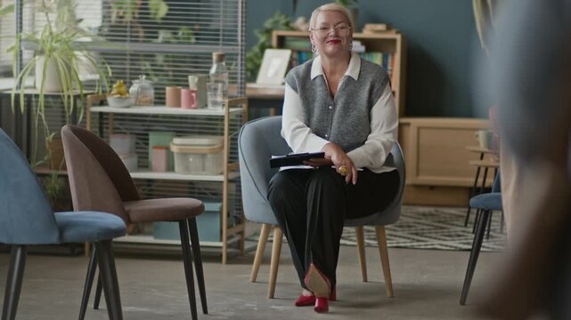 Full shot of friendly mature Caucasian female psychologist welcoming multiethnic group of three students entering cozy modern office and taking seats starting group therapy