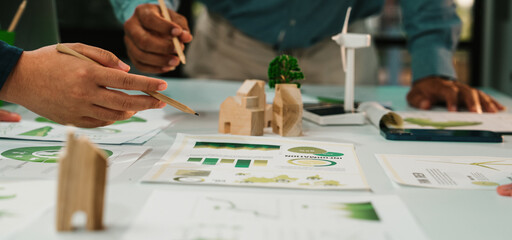 A collaborative team engages in a sustainable design project, analyzing green concepts and innovative ideas with charts and wooden models in a modern office setting. SACTR