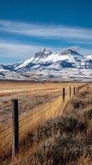 A vertical view of a snowcovered mountain range in wyoming, with a barbed wire fence in the foreground and a blue sky with clouds above, showcasing the natural beauty