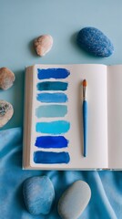 A sketchbook displays blue paint swatches alongside a brush, complemented by pebbles and a cloth, all set against a serene blue backdrop, creating a harmonious artistic scene