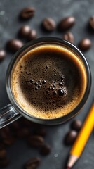 Aromatic coffee in a clear glass mug, surrounded by scattered coffee beans on a textured gray surface, with a yellow pencil adding a pop of color to the scene