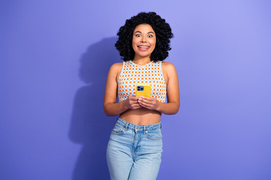 Excited young woman with curly hair using a smartphone against a vibrant purple background showcasing joy and style