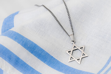A silver-toned Star of David (Magen David) pendant on a chain rests on a softly draped blue and white striped fabric, clearly highlighting the Jewish religious and cultural symbol.
