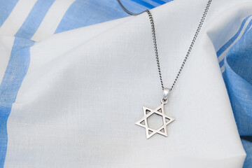 A silver-toned Star of David (Magen David) pendant on a chain rests on a softly draped blue and white striped fabric, clearly highlighting the Jewish religious and cultural symbol.