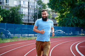A bearded man wearing a light blue shirt jogging on a red track with urban buildings and lush greenery in the background, enjoying a healthy and active lifestyle outdoors.