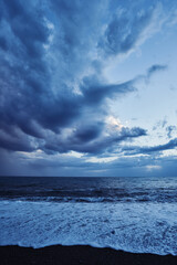 Ocean waves gently crash against the sandy beach under a dramatic sky filled with storm clouds. The serene and somber scene showcases the power and tranquility of nature during twilight.