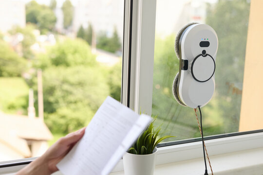 Robotic window cleaner methodically washing glass surface while woman reviewing manual, highlighting smart home technology efficiency