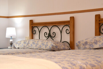A bedroom with two beds decorated in white and blue. The headboards are wrought iron and there is a lamp with the light on.