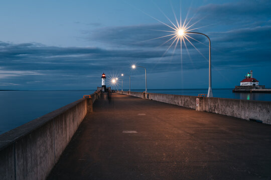Pier leading to the Duluth North Pierhead Lighthouse at dusk on Lake Superior in Duluth, Minnesota, with glowing streetlights, calm water, and the Duluth South Breakwater Lighthouse in the background.