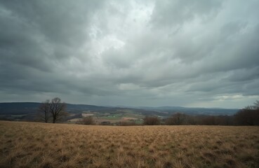 Overcast sky looms above a dry grass field with distant rolling hills and a rural landscape. Bare trees dot the horizon under a moody atmosphere, suggesting an approaching storm.