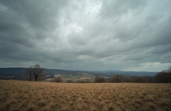 Overcast sky looms above a dry grass field with distant rolling hills and a rural landscape. Bare trees dot the horizon under a moody atmosphere, suggesting an approaching storm.