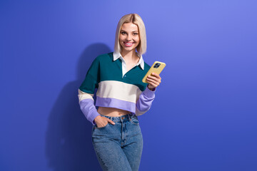 Smiling young blonde woman holding a smartphone in casual stylish outfit against vibrant violet wall