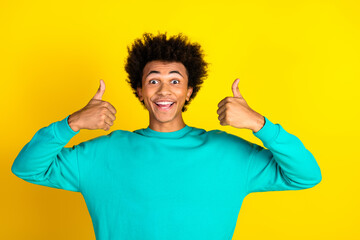 Cheerful young man in a vibrant teal sweatshirt showing thumbs up while smiling brightly against a...